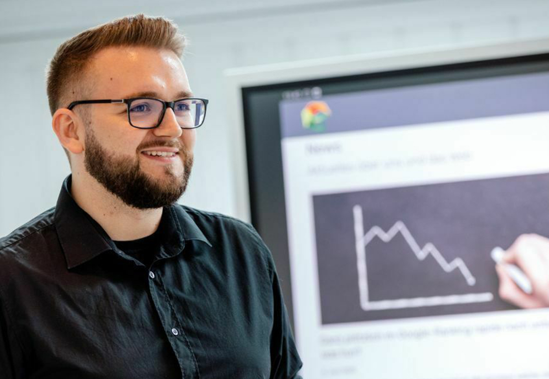 Ein Mann mit Brille und Bart steht vor einem Display und schaut in die Kamera.