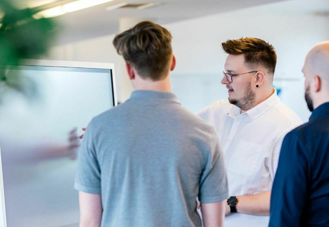 Drei Männer stehen um ein Whiteboard und diskutieren, während sie auf die darauf skizzierten Informationen zeigen.