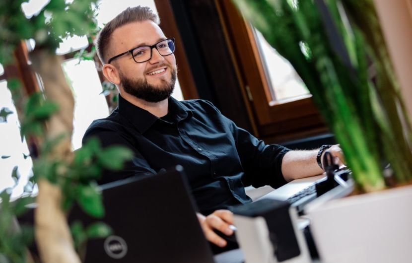 Lächelnder Geschäftsmann sitzt vor Computer und arbeitet, Vordergrund eine Pflanze.