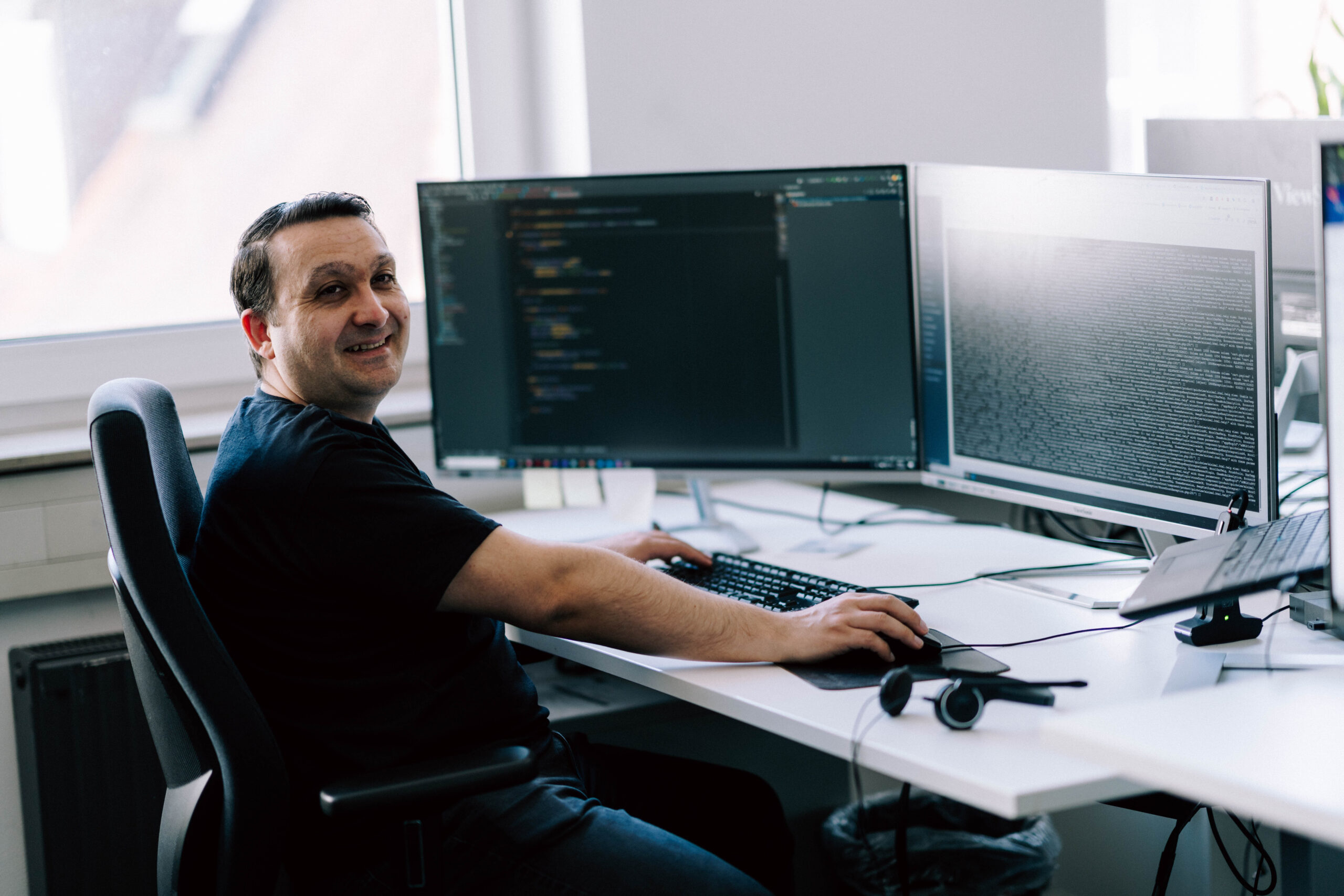 Performance-Optimierung für Ihren Shopware-Shop Das Bild zeigt eine Person, die an einem Schreibtisch vor zwei Monitoren sitzt und am Computer arbeitet. Auf beiden Bildschirmen ist Programmiercode bzw. eine Entwicklungsumgebung zu sehen – vermutlich ein Code-Editor auf dem einen Monitor und ein Terminal oder eine Anwendungsausgabe auf dem anderen.