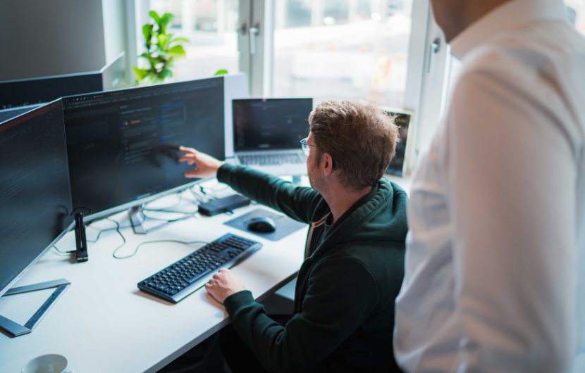 Shopware-Betreuung-betrieb Ein Mann sitzt an einem Schreibtisch vor mehreren Monitoren und zeigt mit der Hand auf den Bildschirm, auf dem Programmcode zu sehen ist. Neben ihm steht eine weitere Person und schaut zu. Der Arbeitsplatz ist modern eingerichtet, mit Tastatur, Maus und Laptop, und befindet sich in einem hellen Büro mit Fenstern im Hintergrund.