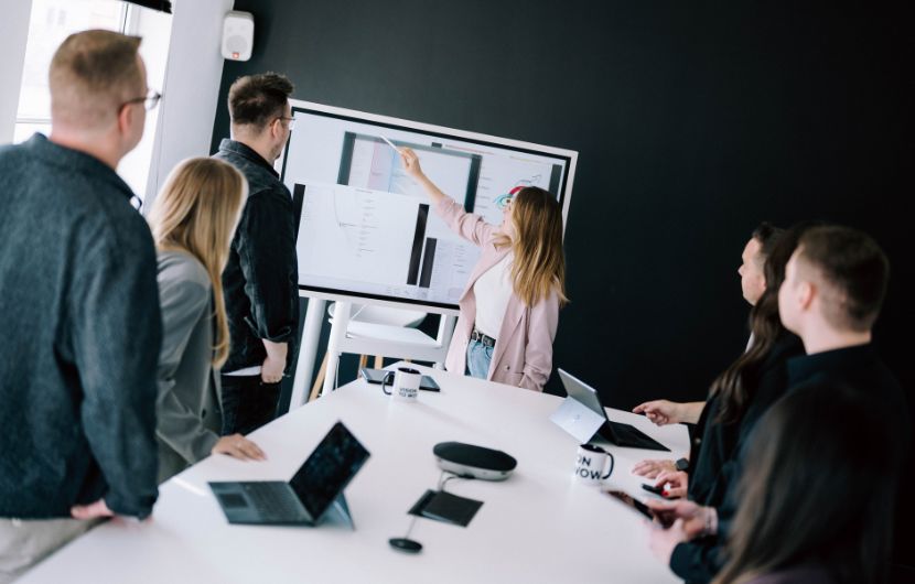 Shopware-schnittstellen-analyse Gruppe von Personen in einem Besprechungsraum schaut auf einen großen Bildschirm, während eine Frau etwas darauf zeigt.