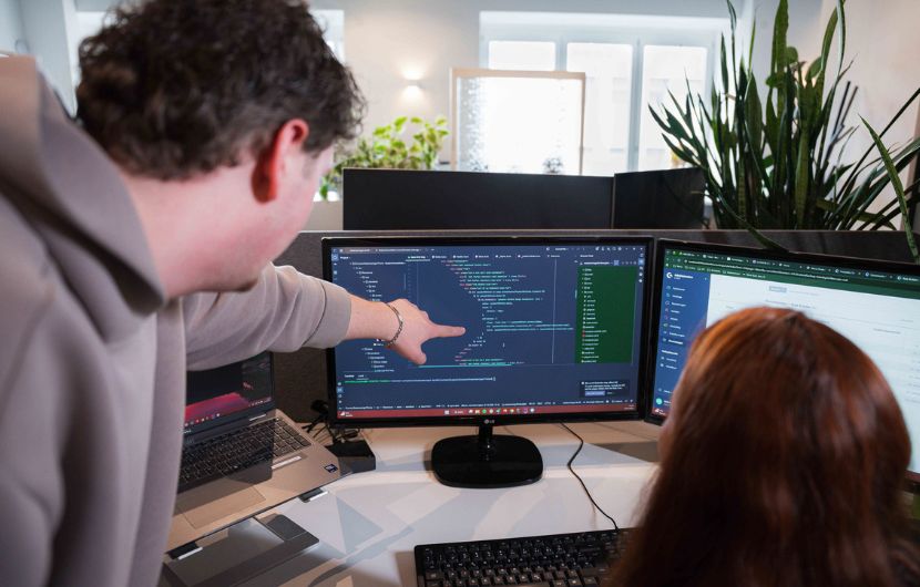 Sicherheitsupdates für Shopware Eine Person beugt sich über einen Schreibtisch und zeigt mit dem Finger auf einen Monitor, auf dem Programmcode zu sehen ist. Eine zweite Person sitzt davor und schaut auf den Bildschirm. Auf dem Schreibtisch stehen eine Tastatur und ein Laptop, während im Hintergrund Pflanzen und weitere Monitore in einem modernen Büro zu sehen sind.