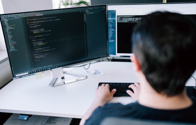 Update-Service-Shopware Person sitzt an einem Schreibtisch und arbeitet an zwei Monitoren mit Programmiercode, während sie auf einer Tastatur tippt.