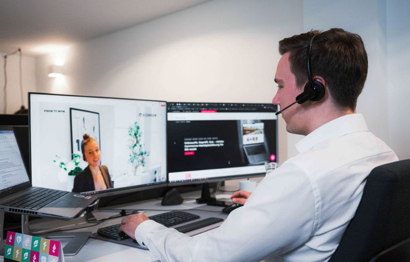 Zukunftssicherheit Ihrers Shopware-projekts Ein Mann sitzt an einem Schreibtisch mit mehreren Monitoren und trägt ein Headset. Auf einem Bildschirm ist ein Videoanruf mit einer Frau zu sehen, auf dem anderen eine Webseite oder Anwendung. Vor ihm stehen Tastatur und Laptop, und er arbeitet in einem modernen, hellen Büro.