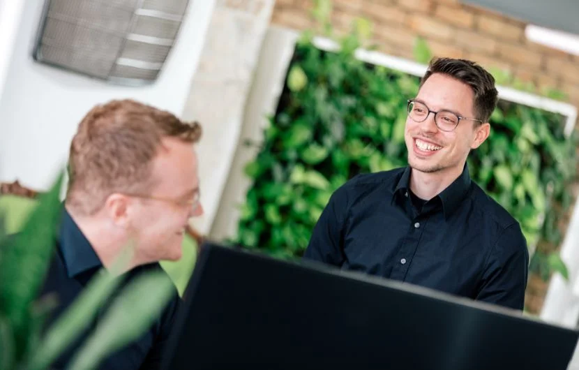 Zwei Online-Shop-Experten in einem modernen Büro mit begrüntem Hintergrund, die sich lächelnd über Suchmaschinenoptimierung unterhalten.