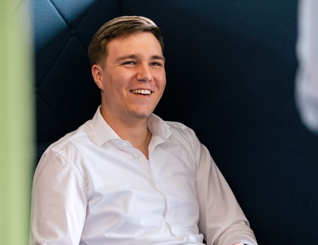 Ein junger Mann sitzt in einem modernen Büro, lächelt und wirkt entspannt, während er an einem E-Commerce-Portalprojekt arbeitet.