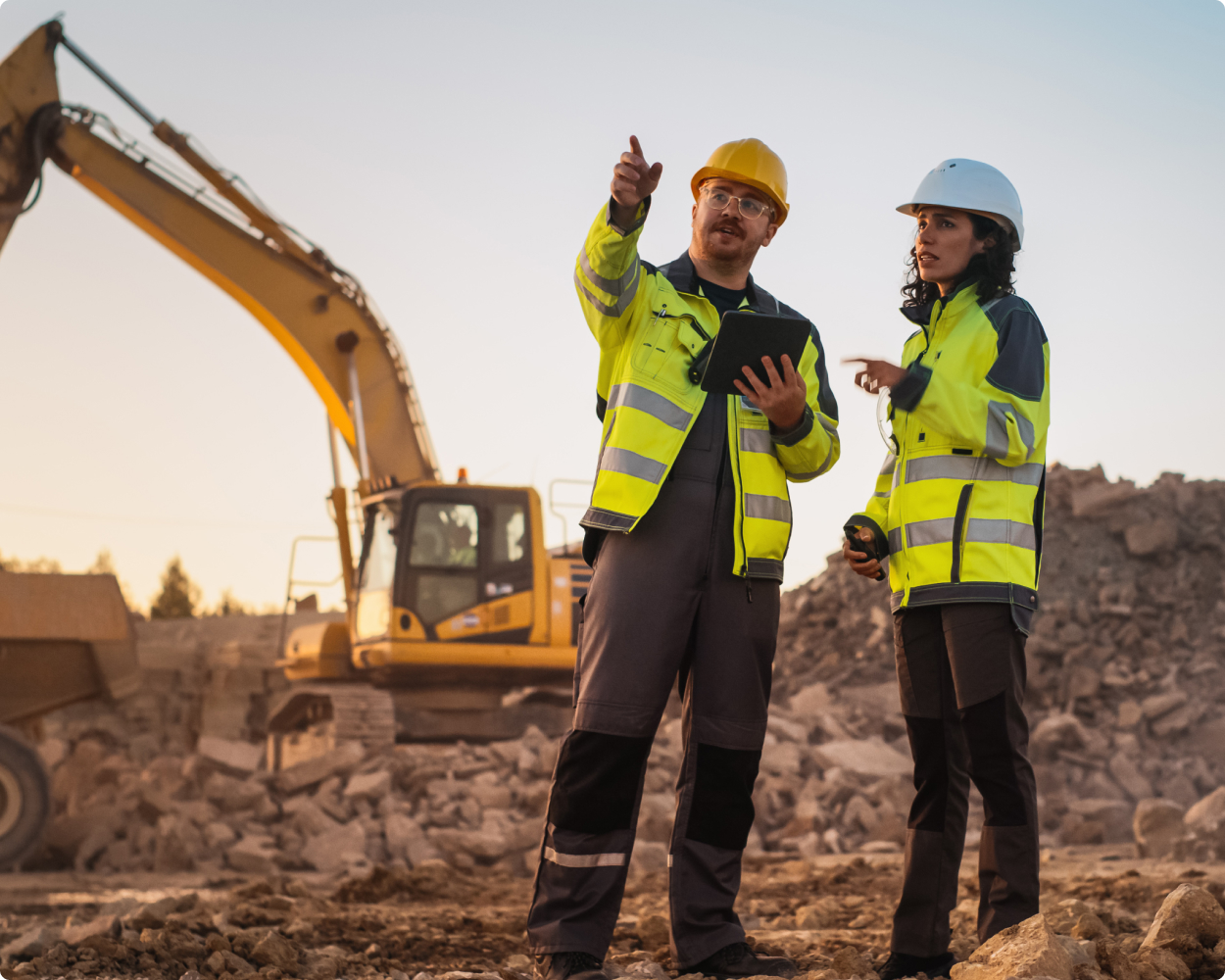 Zwei Bauarbeiter in Warnschutzkleidung von Kübler Workwear und Schutzhelmen stehen auf einer Baustelle mit Schutt und Bagger im Hintergrund. Sie diskutieren und zeigen in eine Richtung, einer hält ein Tablet.