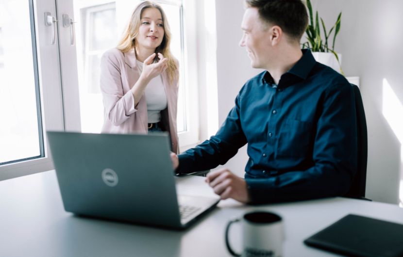 magento-plugins Eine Frau steht neben einem Schreibtisch und spricht mit einem sitzenden Mann, der vor einem Laptop arbeitet. Sie gestikuliert mit der Hand, während er sich ihr zuwendet und zuhört. Auf dem Tisch stehen ein Laptop, eine Tasse und ein Notizbuch. Die Szene spielt in einem hellen Büro mit großen Fenstern.