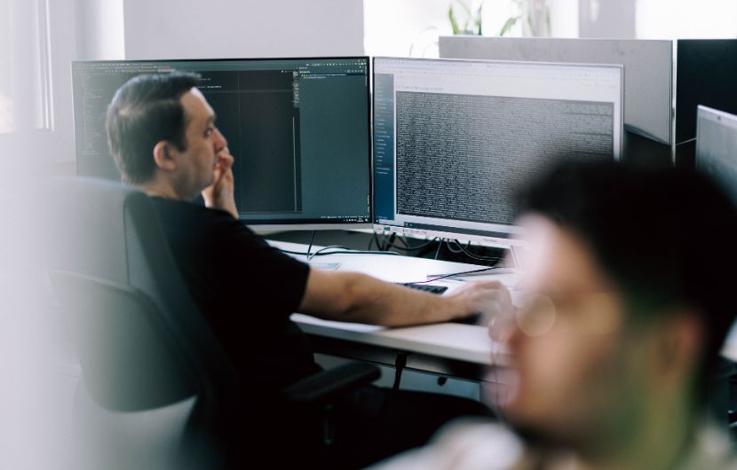 magento-programmierung-qulität Ein Mann sitzt in einem Büro an einem Schreibtisch mit zwei Monitoren und betrachtet konzentriert den angezeigten Programmiercode. Er stützt sein Gesicht mit der Hand, während er mit der anderen die Maus bedient. Im Vordergrund ist eine weitere Person unscharf zu erkennen, was auf eine Arbeitsumgebung mit mehreren Personen hindeutet.