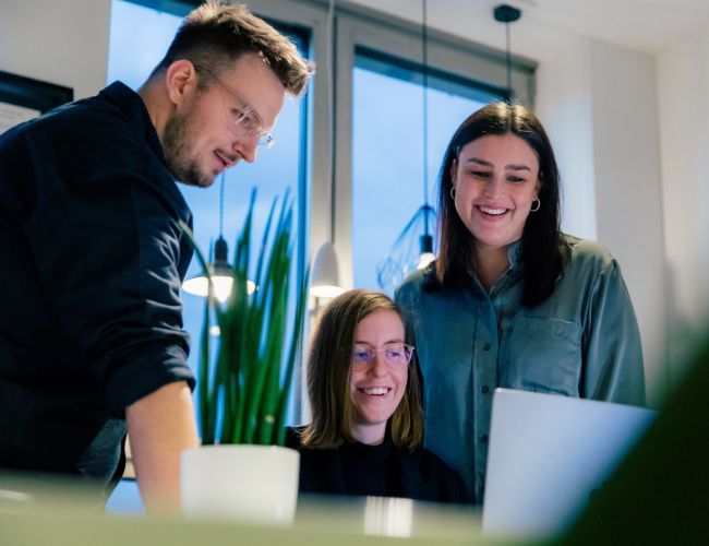 Drei Mitarbeitende der ECONSOR Digitalagentur stehen in einem modernen Büro um einen Laptop und diskutieren strategische Onlineberatung für Unternehmen – individuell und praxisnah für KMU & Konzerne.