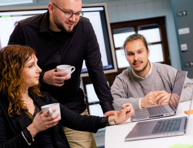 Ein Team aus drei Personen steht zusammen und schaut auf einen Laptop, während sie eine Besprechung zu E-Commerce und Portalentwicklung führen.