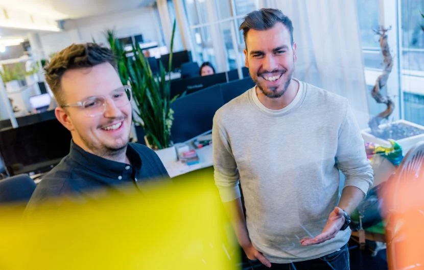 Zwei Mitarbeiter betrachten Post IT's, die an einer durchsichtigen Glaswand befestigt sind und lächeln.