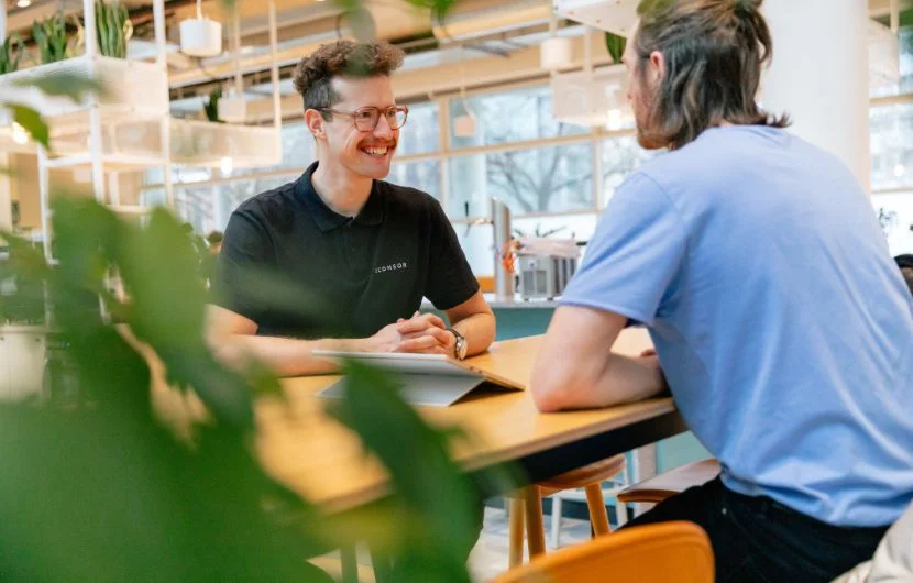 Zwei Mitarbeiter, einer davon lächelt, der andere steht mit dem Rücken zur Kamera sitzen an einem Tisch und unterhalten sich.