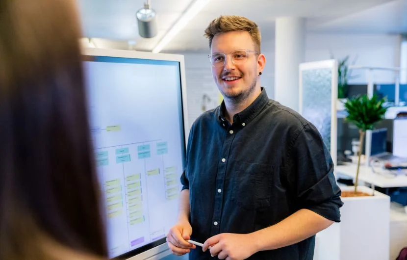 Ein Mitarbeiter lächelt und stellt vor einem Whiteboard eine Präsentation vor.