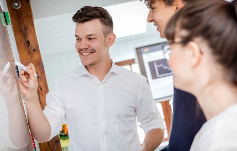 Ein junger Mann in weißem Hemd erstellt zusammen mit zwei Kolleg*innen eine Social-Media-Strategie an einem Whiteboard – symbolisiert die Zusammenarbeit in einer Social-Media-Agentur, die auf effektives Social-Media-Marketing setzt.