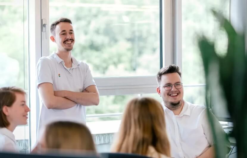 In einer Besprechung einer Webdesign-Agentur unterhalten sich mehrere junge Mitarbeitende gut gelaunt in einem modernen, hellen Büro mit großen Fenstern.