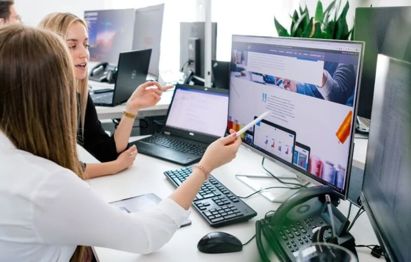 In einer Besprechung einer Webdesign-Agentur besprechen zwei Mitarbeiterinnen gemeinsam die Gestaltung einer Webseite an einem großen Bildschirm.