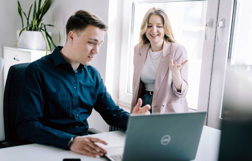 SEO-Analyse im Team für mehr Website-Effizienz Zwei Marketing-Profis sitzen an einem Schreibtisch und besprechen angeregt Inhalte auf einem Laptop. Die Frau gestikuliert freundlich, während der Mann auf den Bildschirm schaut. Die Szene vermittelt eine kooperative Arbeitsatmosphäre in einer Agentur bei einem SEO-Audit.
