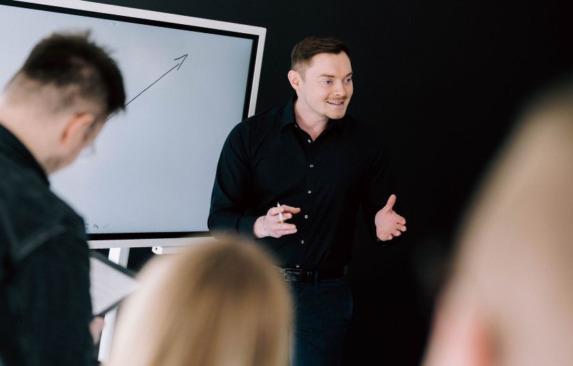 Ein Mann in dunkler Kleidung hält eine lebendige Präsentation vor einem Bildschirm mit einem aufsteigenden Pfeil. Vor ihm sitzen mehrere Zuhörer. Die Szene zeigt eine professionelle Umgebung für ein SEO-Audit oder eine Strategiebesprechung zur Effizienzsteigerung.