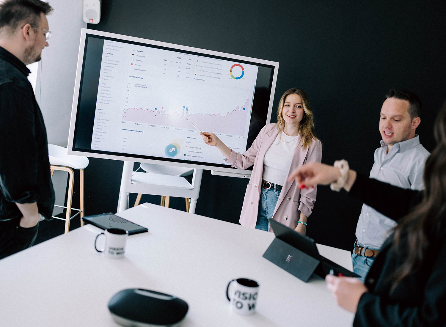 Agenturteam im modernen Konferenzraum, Frau zeigt Diagramme auf großem Display zur Online‑Strategie
