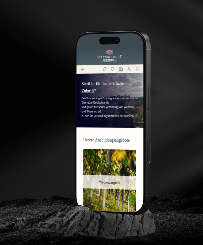 Ein Smartphone steht hochkant auf einem düsteren und unebenem Stein. Auf dem Bildschirm des Smartphones ist der Staatsweingut Freiburg-Shop abgebildet.