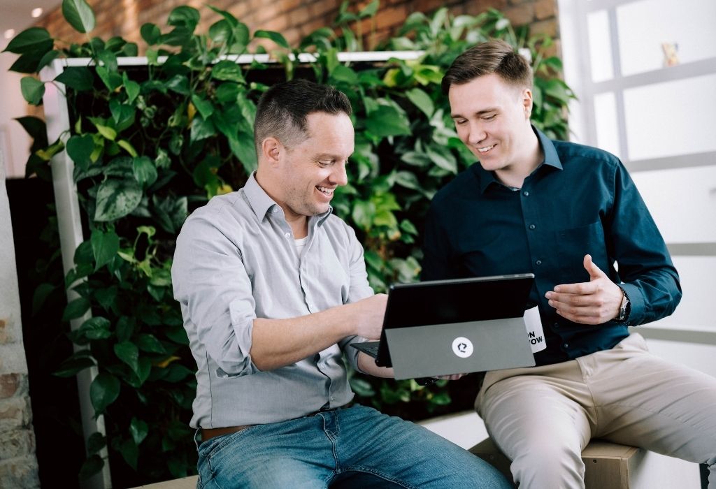 Zwei Männer sitzen vor einem geöffneten Laptop, betrachten gemeinsam Inhalte und führen eine sachliche Besprechung in einem Bürobereich mit grüner Pflanzenwand.