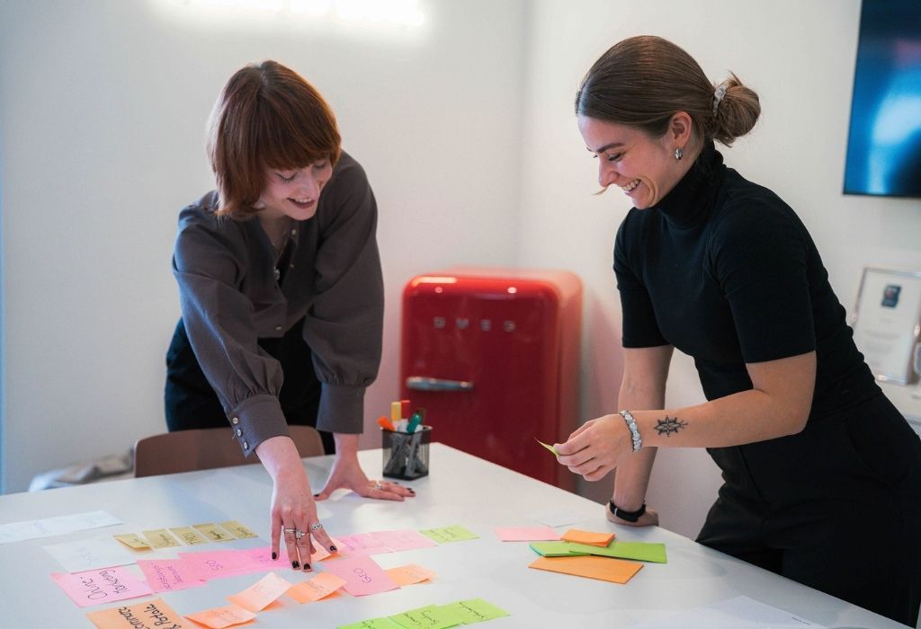 Zwei Mitarbeiterinnen arbeiten an einem Tisch voller farbiger Post-its, sortieren Notizen und entwickeln gemeinsam Ideen in einem modernen Besprechungsraum.