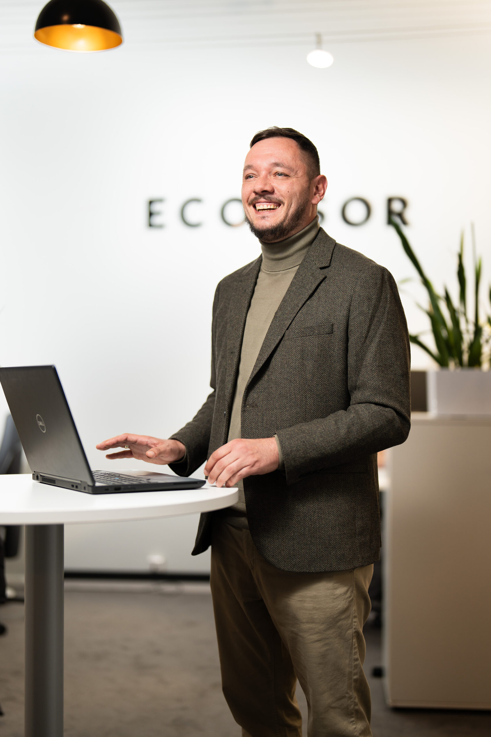Freundlicher Mitarbeiter Ein lächelnder Mann arbeitet an einem Laptop an einem weißen Stehtisch in einem modernen Büro. Er trägt Rollkragenpullover, Sakko und helle Hose, im Hintergrund sind Firmenname und Pflanzen zu sehen.