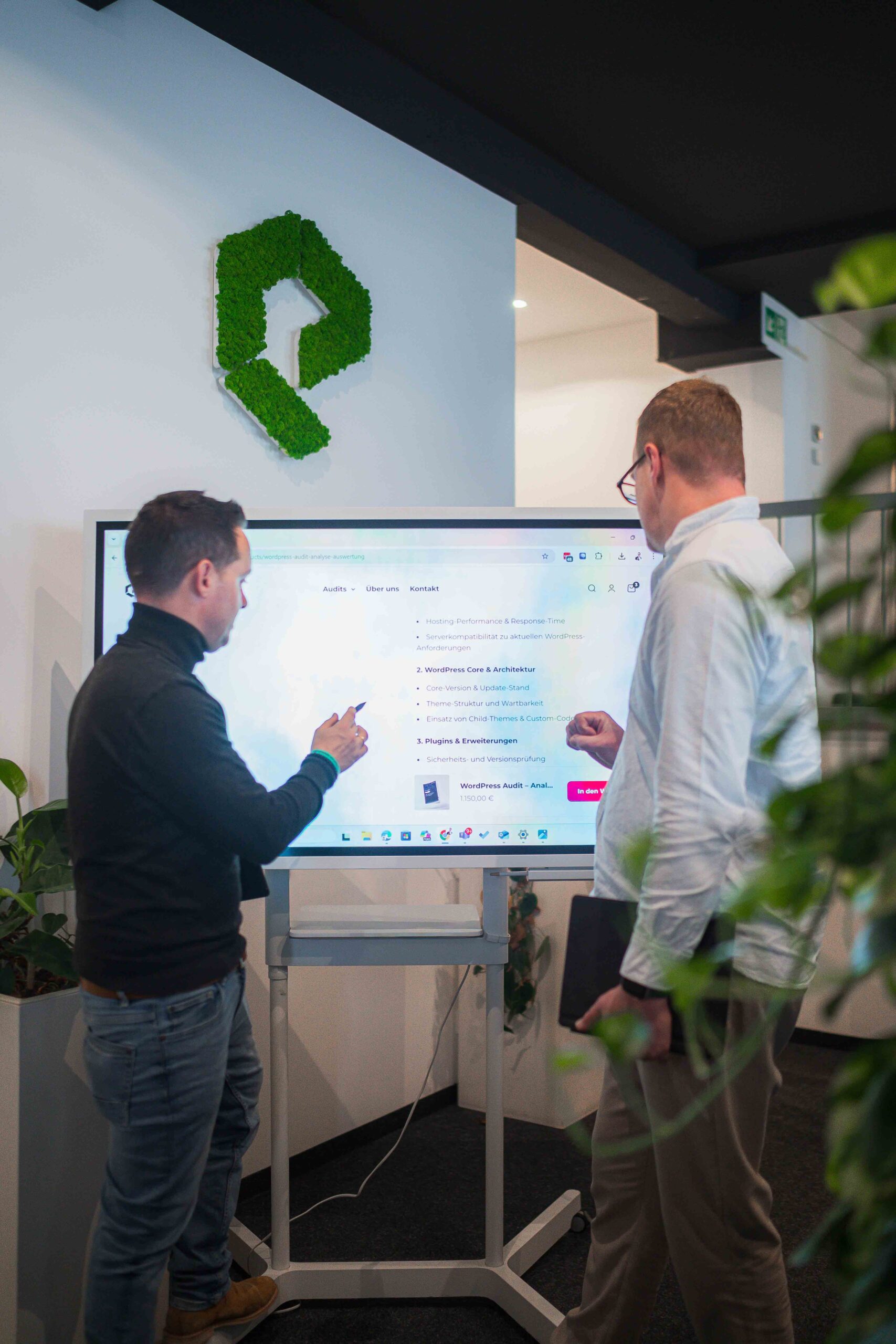 Gemeinsame Analyse am Touchscreen Zwei Personen stehen vor einem interaktiven Bildschirm und besprechen Inhalte, umgeben von modernen Büroelementen und Pflanzen.