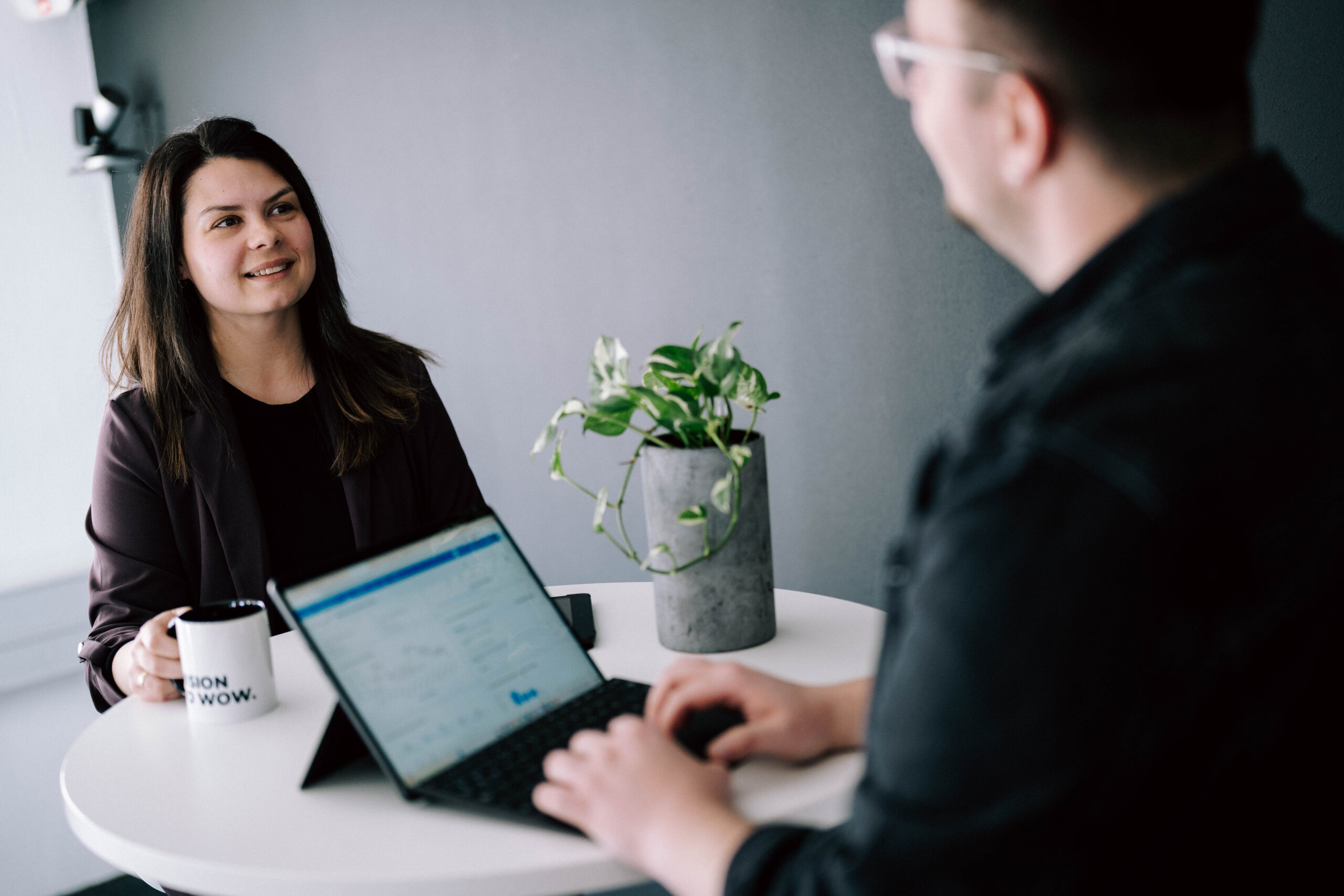 Ein Gespräch zwischen zwei Personen in einem modernen Büro. Eine Frau hält eine Tasse mit dem Schriftzug „VISION WOW“ in der Hand, während sie einem Mann gegenüber sitzt, der auf ein Tablet schaut. Auf dem Tisch steht eine Pflanze in einem grauen Topf.