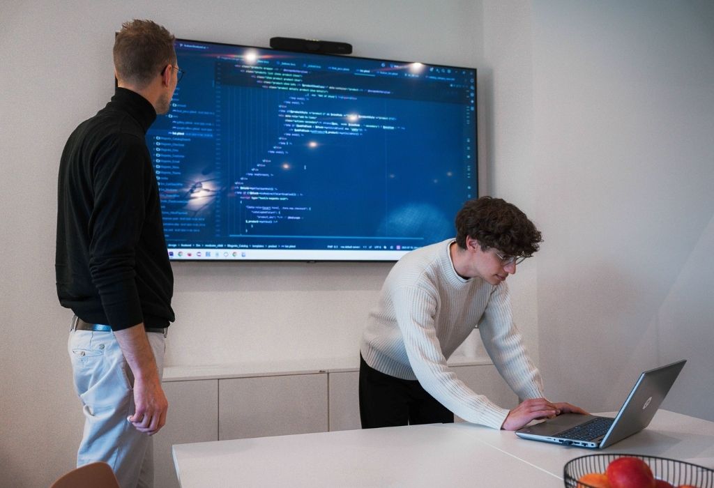 Teamarbeit bei der Code-Durchsicht im Büro Zwei Kollegen in einem Meeting, die Code auf einem großen Bildschirm und einem Laptop besprechen.