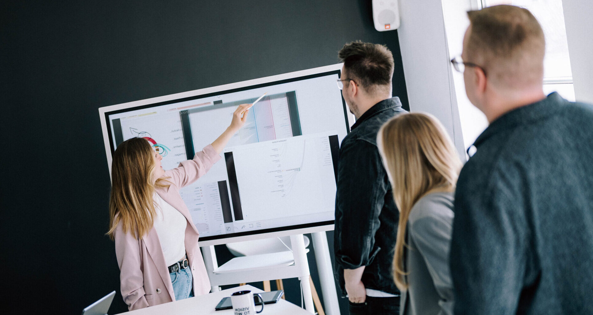 Eine Gruppe von vier Personen bespricht auf einem modernen Touchscreen-Monitor eine Präsentation. Eine Person zeigt mit einem Stift auf die Bildschirmansicht, während die anderen aufmerksam zuhören. Der Raum ist modern gestaltet mit dunklen Wänden und einem Schreibtisch, auf dem ein Laptop und eine Tasse stehen.