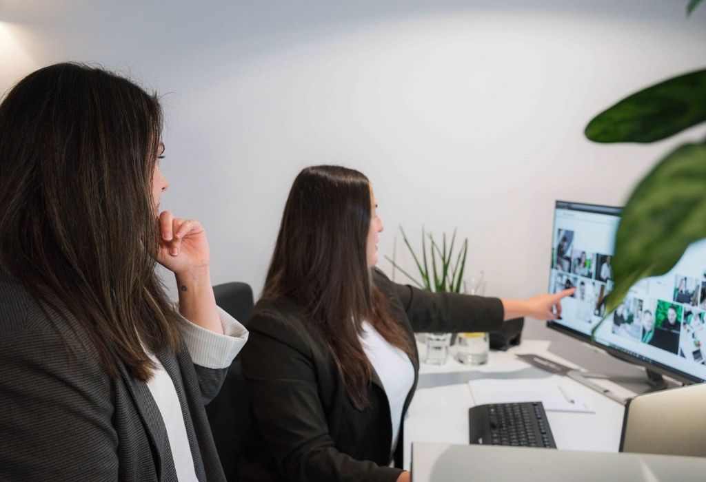 Zwei Frauen sitzen im Büro vor einem Monitor, sprechen über die angezeigten Inhalte und eine zeigt mit dem Finger auf eine Bildübersicht auf dem Bildschirm.