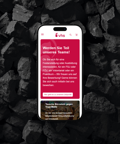 Ein Smartphone liegt flach auf einer dunklen, steinigen Oberfläche. Auf dem Bildschirm des Smartphones ist die vhs-Website abgebildet.