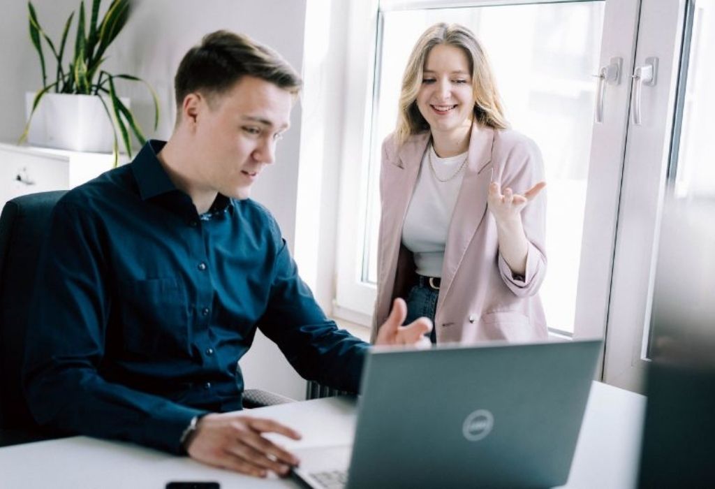 Zwei Marketing-Profis sitzen an einem Schreibtisch und besprechen angeregt Inhalte auf einem Laptop. Die Frau gestikuliert freundlich, während der Mann auf den Bildschirm schaut. Die Szene vermittelt eine kooperative Arbeitsatmosphäre in einer Agentur bei einem SEO-Audit.