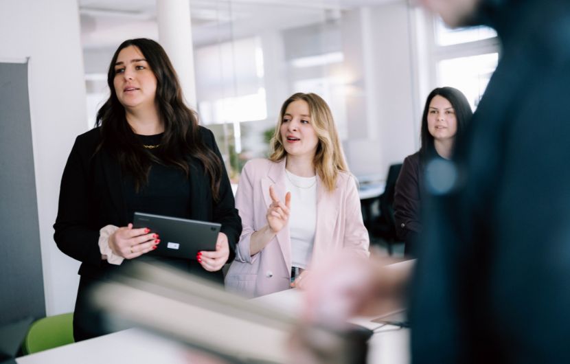 Performance-Optimierung.coversion-analyse Geschäftsfrauen im Büro arbeiten gemeinsam an einem Tablet, Teammeeting und Zusammenarbeit im modernen Unternehmen, während eine Kollegin zuschaut und eine Person im Vordergrund unscharf ist.