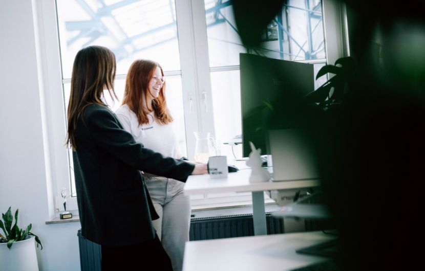 Shopware-SLA-Optionen Das Bild zeigt zwei Frauen, die gemeinsam an einem Stehschreibtisch in einem hellen Büro arbeiten. Vor ihnen stehen ein oder mehrere Monitore, auf die sie schauen, während eine der beiden offenbar etwas erklärt oder zeigt.