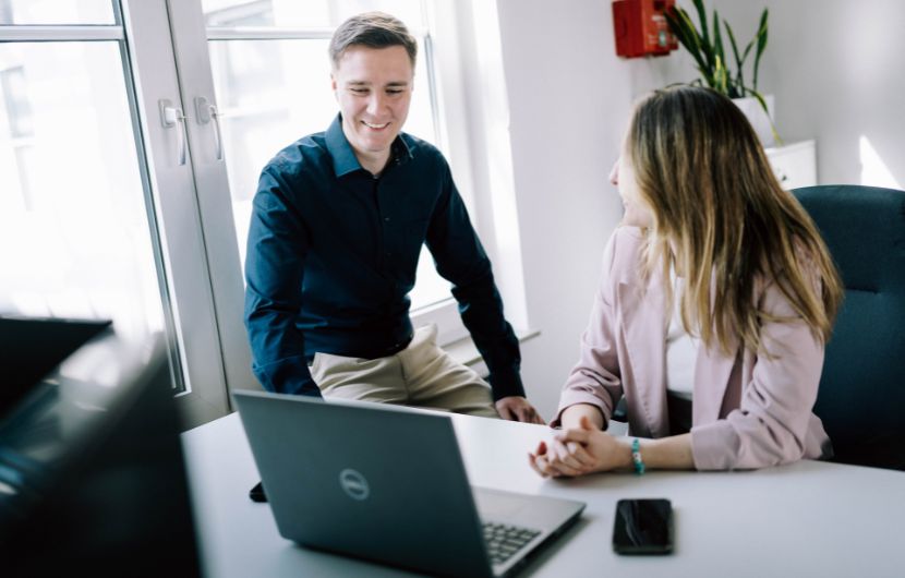 Shopware-Support-Prozesse Das Bild zeigt zwei Personen, die gemeinsam an einem Schreibtisch sitzen und sich unterhalten. Vor ihnen steht ein Laptop, der geöffnet ist, sowie ein Smartphone auf dem Tisch.