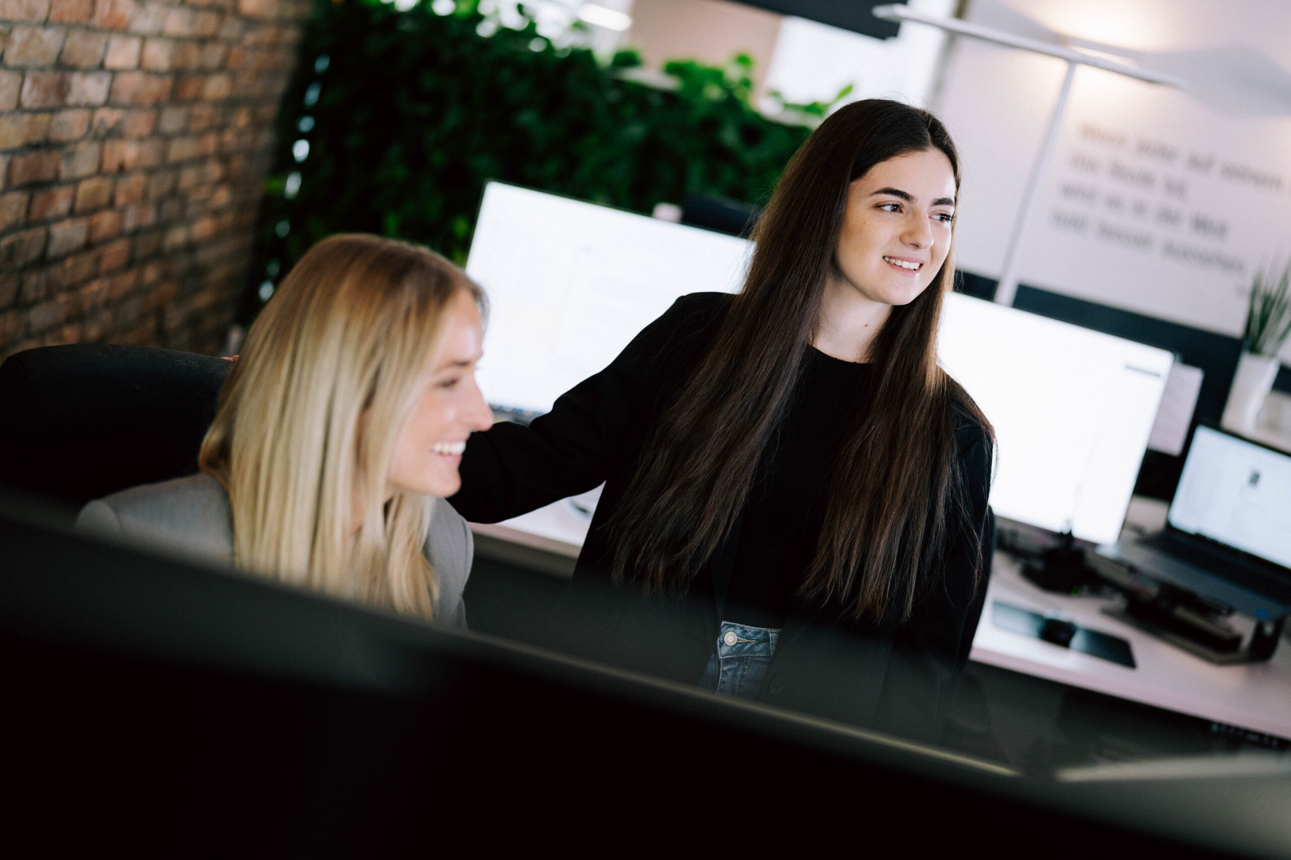 Warum E-Commerce-Projekte nicht an Features scheitern, sondern am Betrieb Zwei Frauen in einem modernen Büro: Eine stehende Frau legt lächelnd ihre Hand auf die Schulter einer sitzenden Kollegin, während sie sich unterhalten. Im Hintergrund sind Computerbildschirme zu sehen.