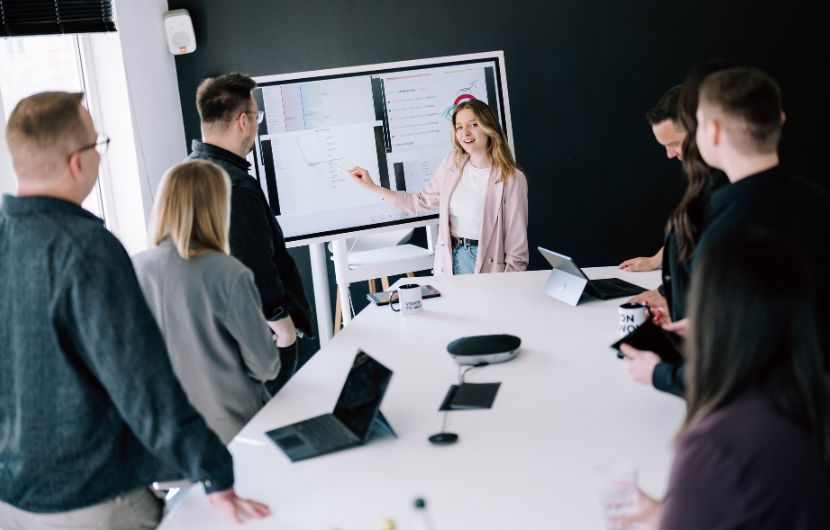 Magento_Architektur_Konzept Teammeeting im Büro – Mitarbeiter stehen um einen Konferenztisch und verfolgen eine Präsentation auf einem großen Bildschirm, während eine Kollegin Inhalte erklärt