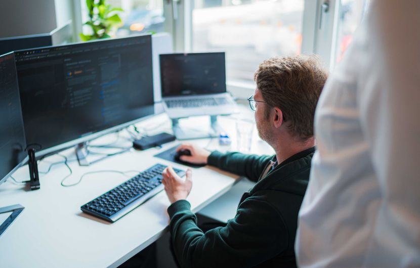 Magento_vorgehen Eine Person sitzt an einem Schreibtisch vor zwei Monitoren und arbeitet an Programmcode. Neben der Tastatur liegt ein Smartphone, im Hintergrund steht ein Laptop. Eine zweite Person steht daneben und schaut zu