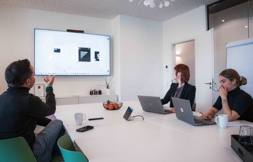 magento-Analyse_Audit Teammeeting im modernen Büro – drei Personen arbeiten gemeinsam an Laptops und verfolgen eine Präsentation auf einem großen Bildschirm während einer Besprechung am Konferenztisch
