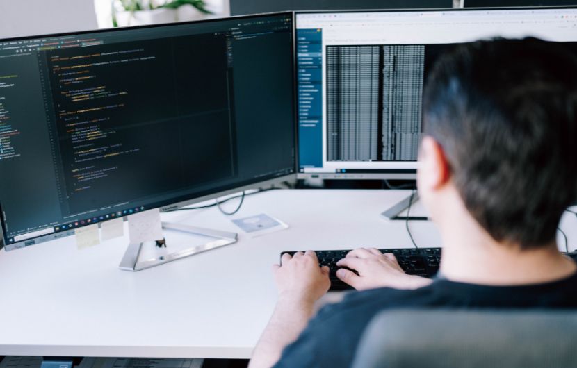 magento_strucktur Eine Person sitzt am Schreibtisch und programmiert an zwei Monitoren – auf einem Bildschirm ist Code zu sehen, auf dem anderen Daten/Logs.
