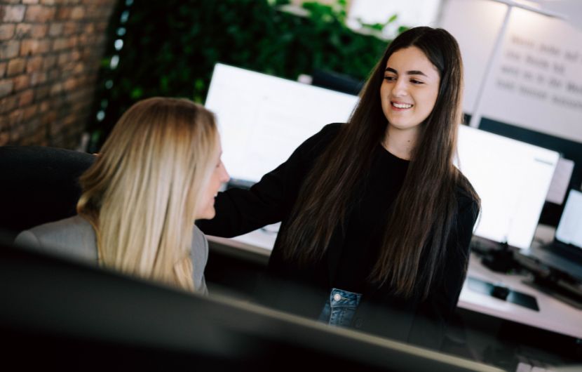 Zwei Frauen in einem modernen Büro: Eine stehende Frau legt lächelnd ihre Hand auf die Schulter einer sitzenden Kollegin, während sie sich unterhalten. Im Hintergrund sind Computerbildschirme und Pflanzen zu sehen.