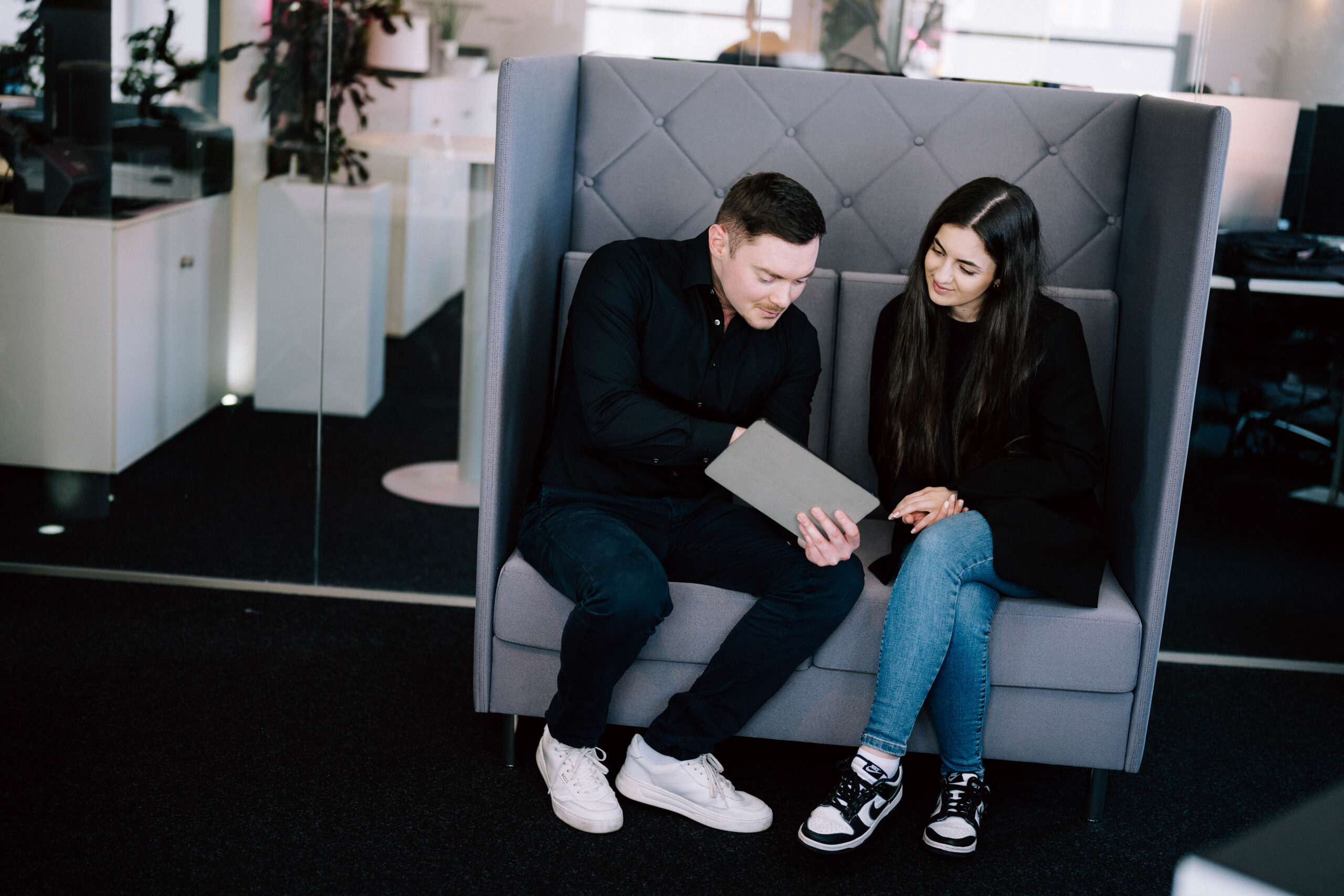 Digitale Services statt Website-Funktionen Zwei Personen sitzen in einer modernen Büroumgebung auf einem grauen Sofa und betrachten gemeinsam ein Tablet.