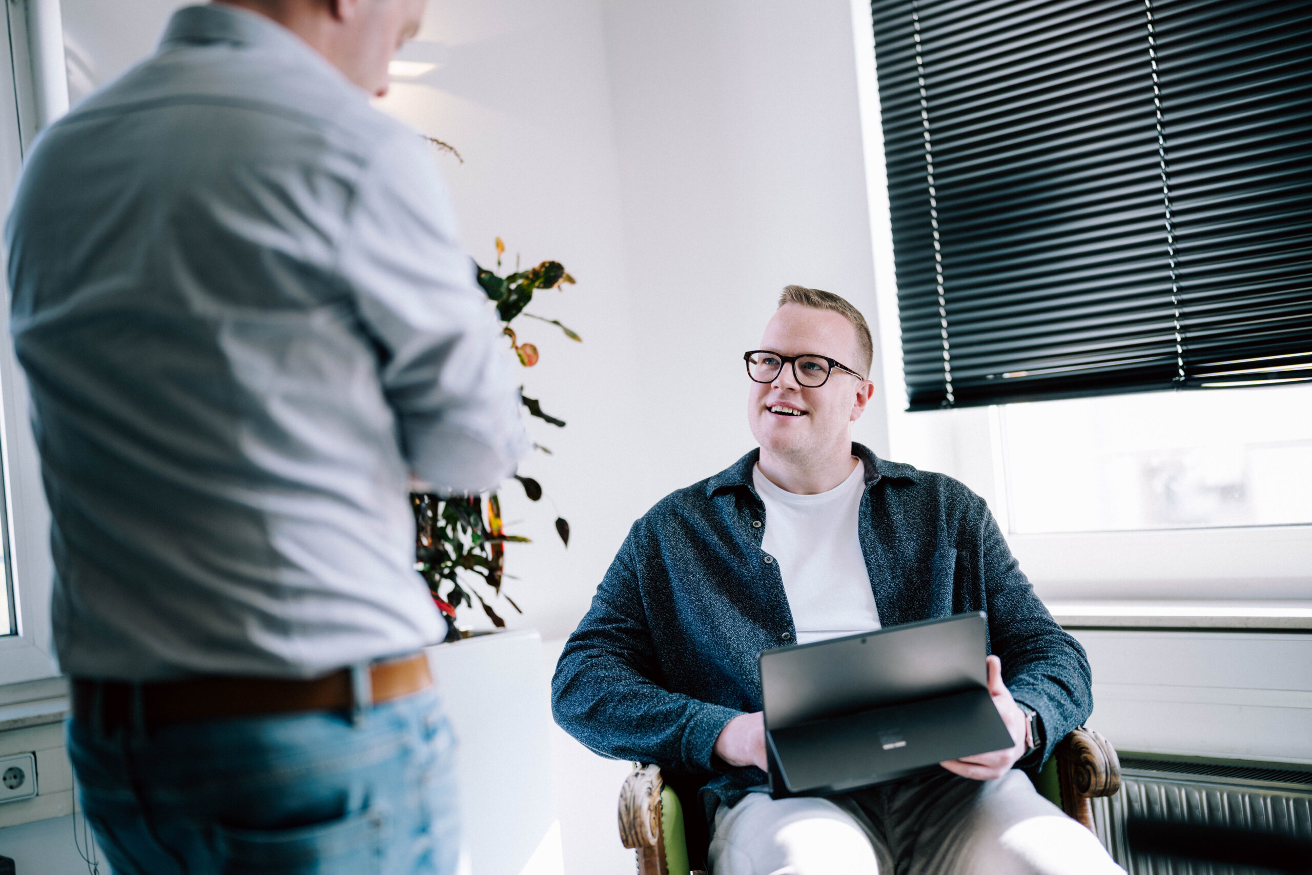 KI und Internationalisierung Zwei Personen im hellen Büro bei einem Beratungsgespräch. Eine Person sitzt mit Laptop im Sessel und blickt lächelnd zur stehenden Person.