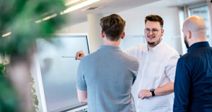 Drei Männer stehen vor einem Smartboard. Der Mann in der Mitte zeigt auf das Board.