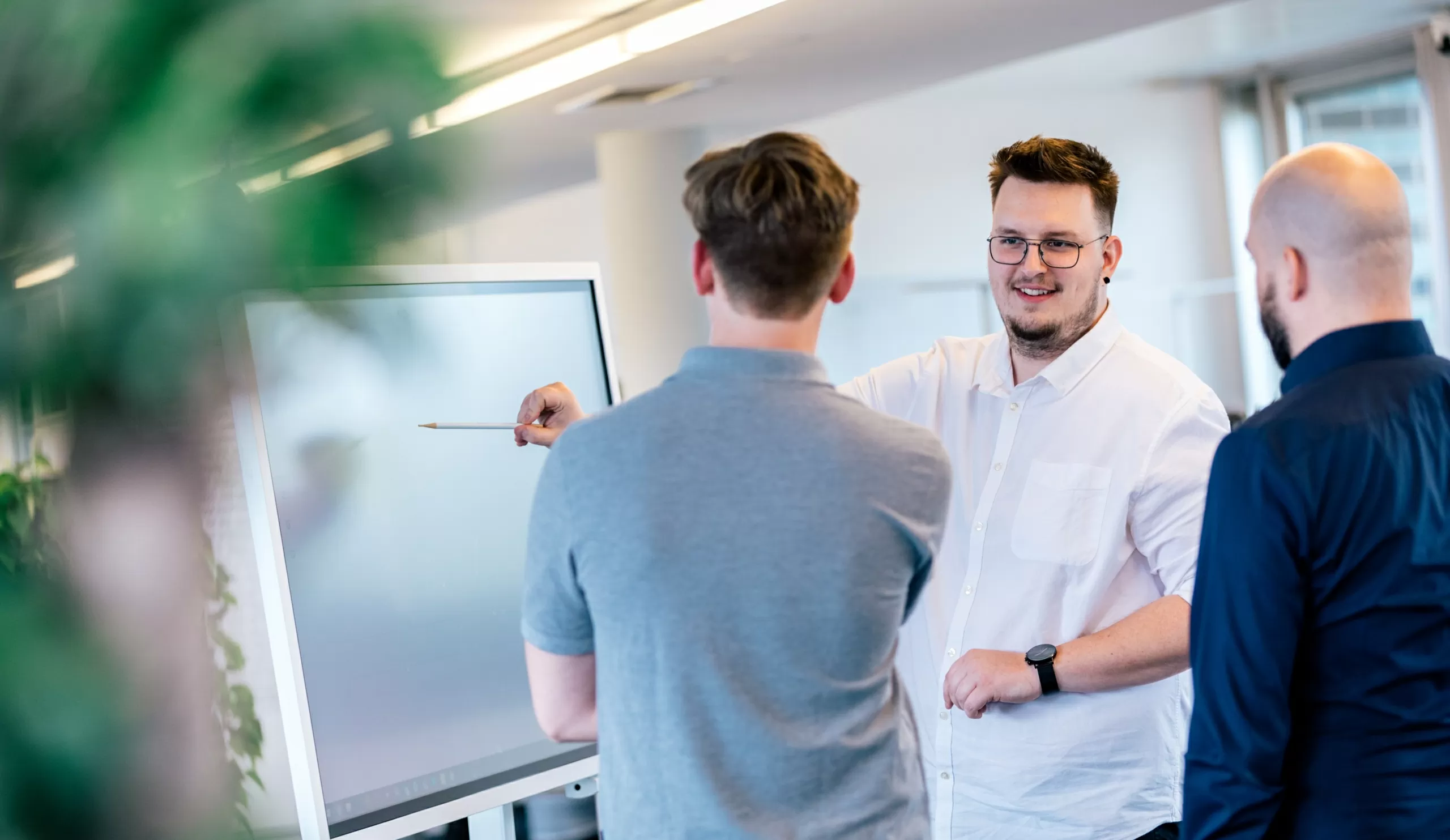 Drei Männer stehen vor einem Smartboard. Der Mann in der Mitte zeigt auf das Board.