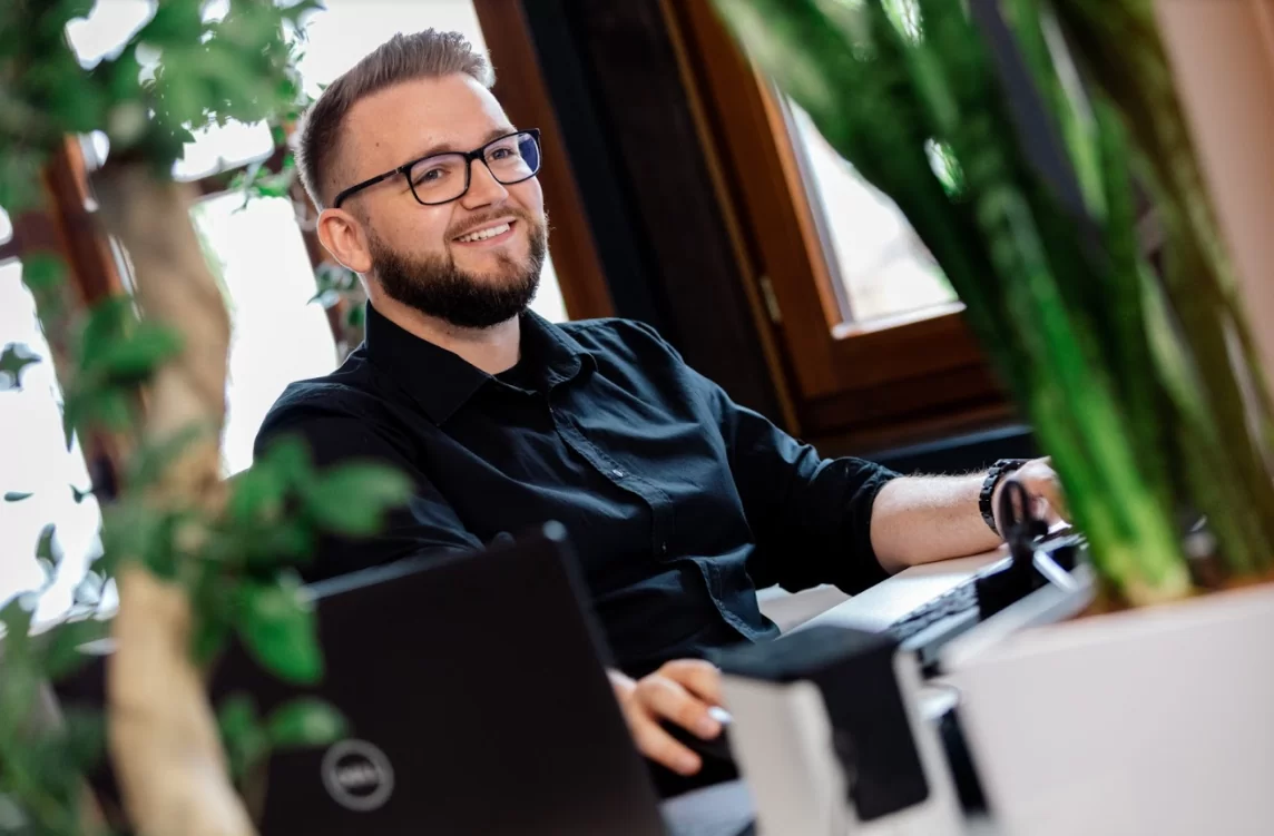 Lächelnder Geschäftsmann sitzt vor Computer und arbeitet, Vordergrund eine Pflanze.